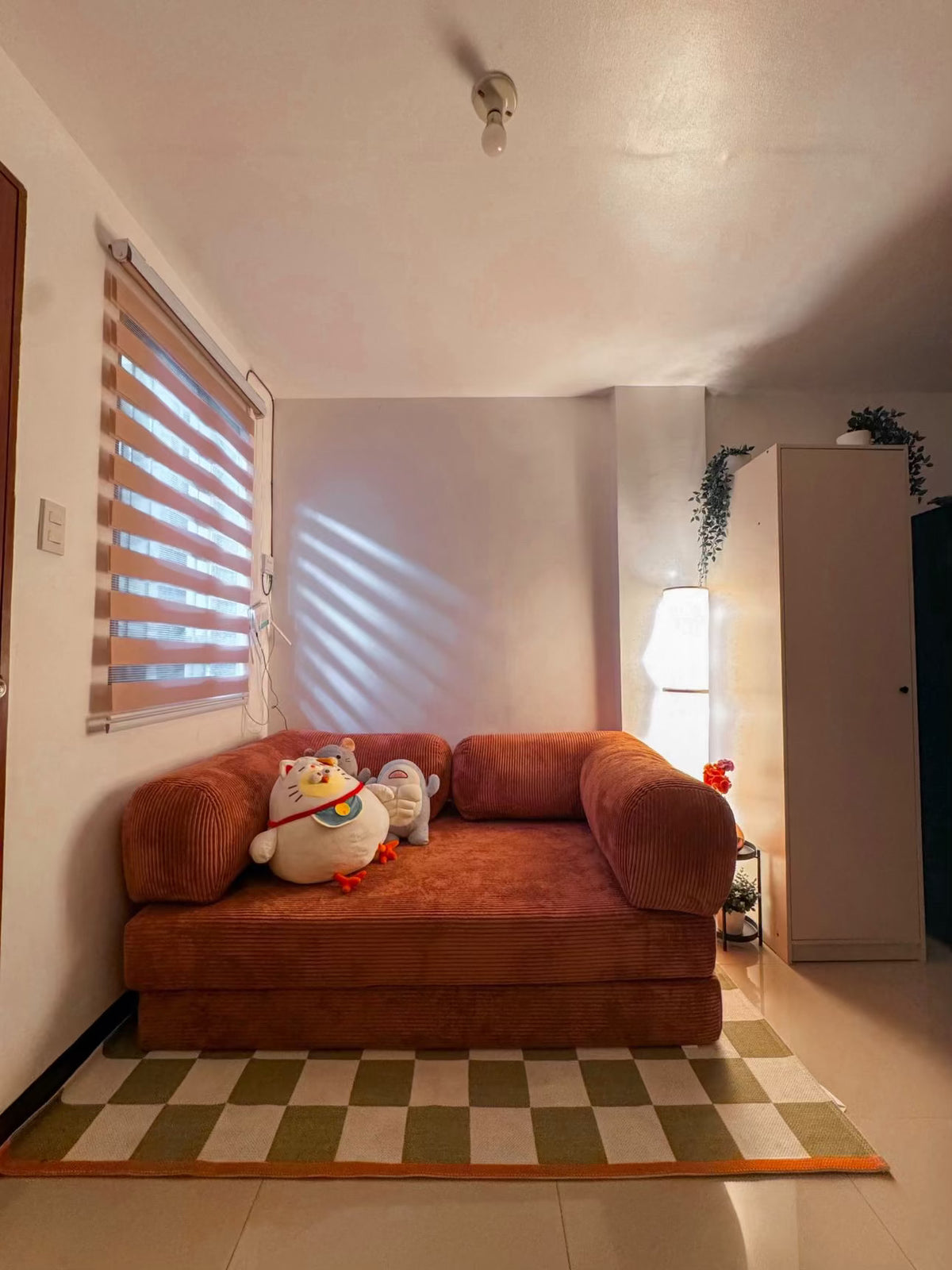 Cozy living room with a rust orange Teddy sofa, playful plush toys, checkered green and white rug, and warm ambient lighting from Home Hues furniture